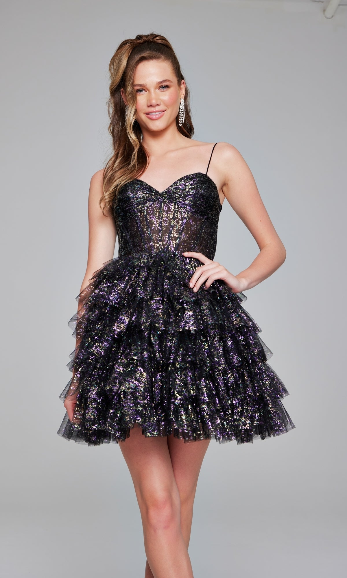 A woman stands smiling against a plain background, wearing the Short Black Glitter Hoco Dress: Jovani 39654, featuring a sparkly finish and a layered ruffle skirt.